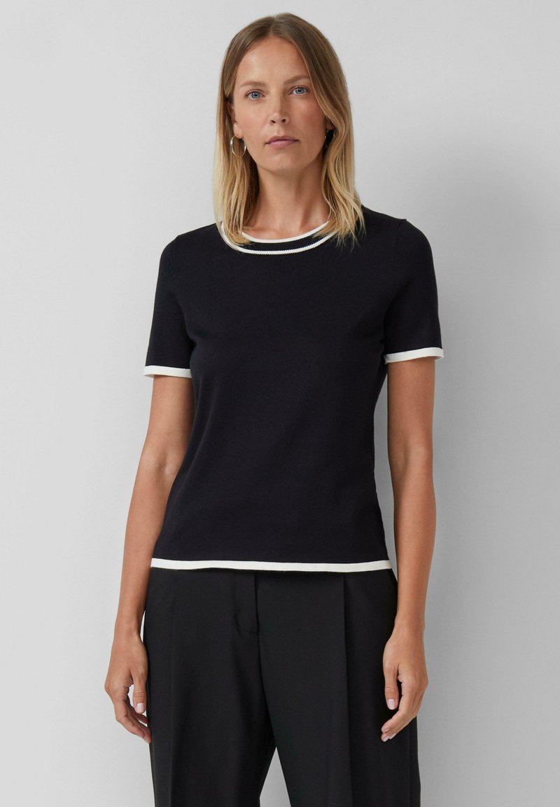 Femme portant un haut noir à manches courtes avec un bord blanc au col, aux manches et à l'ourlet, associé à un pantalon noir, se tenant contre un fond uni.