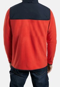 Man in een rode fleecejack met een donkerblauwe panel op de bovenrug, in combinatie met donkerblauwe jeans, bekeken van achteren.