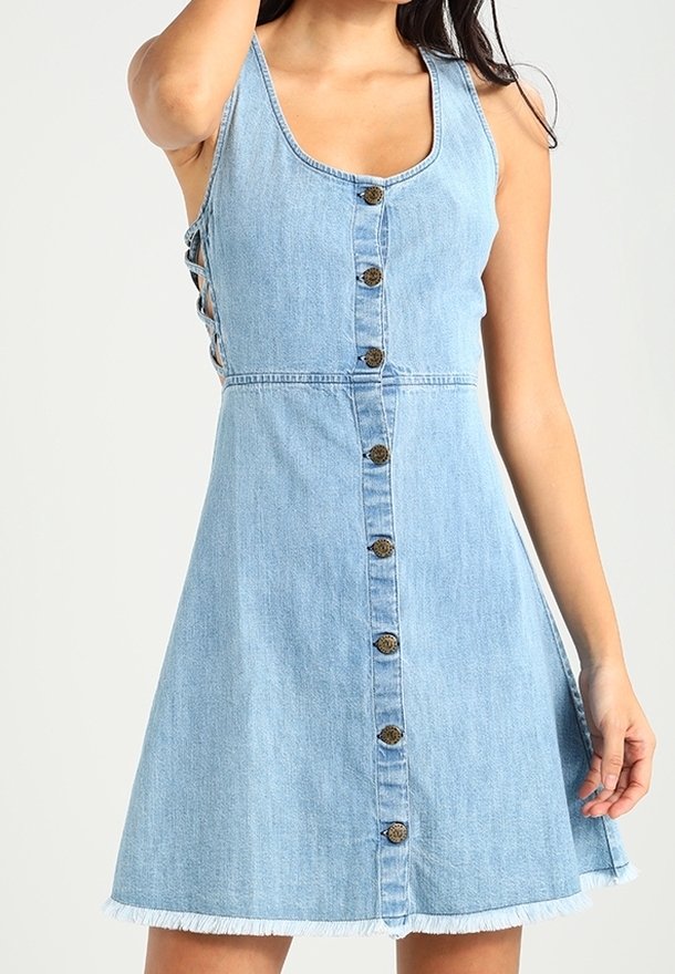 Robe en denim bleu clair sans manches, avec fermeture à boutons sur le devant et ourlet à franges. Présente des détails de laçage sur les côtés pour une texture ajoutée.