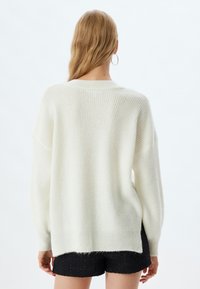 Hellecremefarbene Sweater mit gerippter Textur, überschnittenen Schultern und seitlichen Schlitzen. Das Design ist locker geschnitten und verfügt über einen weiten Ausschnitt und lange Ärmel.