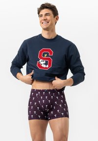 Homme souriant levant un sweat-shirt bleu marine avec un "S" rouge et un graphique de Snoopy, portant un boxer bleu marine à motif de petits personnages Snoopy et Woodstock.