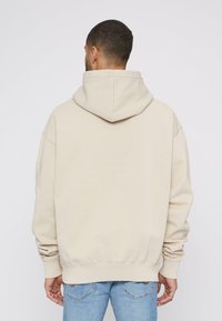 Beige hættetrøje med stor hætte, dropped skuldre og ribbede manchetter. Glat stofstruktur; båret med lyseblå jeans.