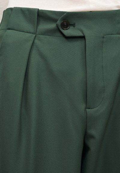 Σκούρο πράσινο παντελόνι tailored με μπροστινό μαύρο κουμπί και πιέτες, φορεμένο με ανοιχτό μπεζ τοπ.