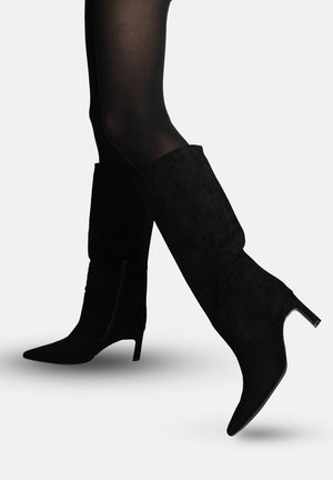 Ben i gennemsigtige sorte strømpebukser iført knælange sorte ruskindsstøvler med spidse tæer og slanke midhøje hæle, den ene fod let løftet.