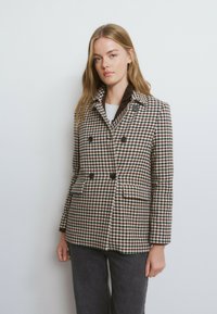Abrigo cruzado estilo peacoat en un patrón de pata de gallo marrón y crema, con dos bolsillos delanteros y botones oscuros; tejido de lana texturizada.