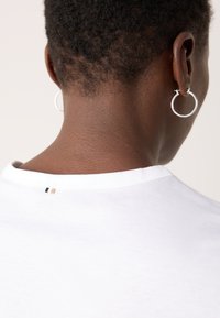 White cotton t-shirt with a round neckline, featuring a small black and gold tag at the back and a silver hoop earring.