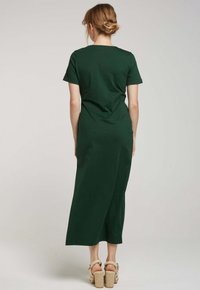 Robe maxi vert foncé à manches courtes avec une silhouette droite, un col rond et une texture subtile ; portée avec des sandales à talons beiges.