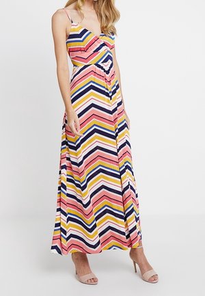 Vrouw draagt een mouwloze maxidress met meerkleurige chevronstrepen en spaghettibandjes, gecombineerd met beige sandalen met hoge hakken.