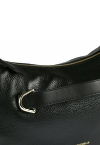 Schwarze Ledertasche mit strukturierter Oberfläche, goldfarbenem Reißverschluss, schwarzem Riemen, der mit einer silbernen Metallschnalle befestigt ist, und teilweise sichtbarem goldfarbenem Markenlogo.