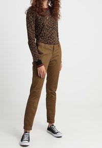 T-shirt marron à manches longues avec un motif floral, associé à un pantalon beige retroussé et des baskets noires. Comprend des poignets contrastés noirs et rouges.