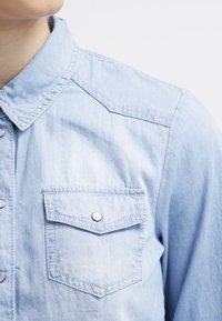 Light blue denim shirt with a button-up front, a collared neck, and one chest pocket featuring a snap closure. Soft texture, casual design.