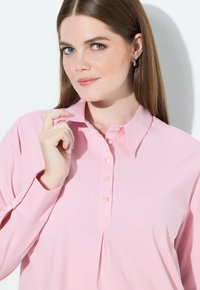 Camicia rosa chiaro con bottoni, colletto appuntito, bottoni perlacei e maniche lunghe, con una piega frontale per un dettaglio aggiuntivo.