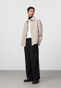 Beige jasje met grote voorzakken, een wit shirt eronder, zwarte broek en zwarte schoenen. Glad materiaal met een getailleerde pasvorm.