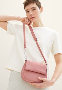 Borsa a tracolla rosa con una texture liscia, dotata di pattina pieghevole, cinturino regolabile e un accentato a catena. Modella che tiene la borsa.