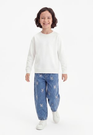 Enfant souriant et marchant, portant un t-shirt blanc à manches longues, un jean bleu avec des imprimés licornes et des baskets blanches sur un fond clair.