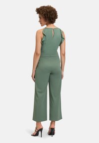 Une combinaison verte dotée d'un buste ajusté, de bretelles volantées, d'une découpe en trou de serrure dans le dos et de pantalons à jambes larges, fabriquée en tissu lisse. Sandales à talons noires.