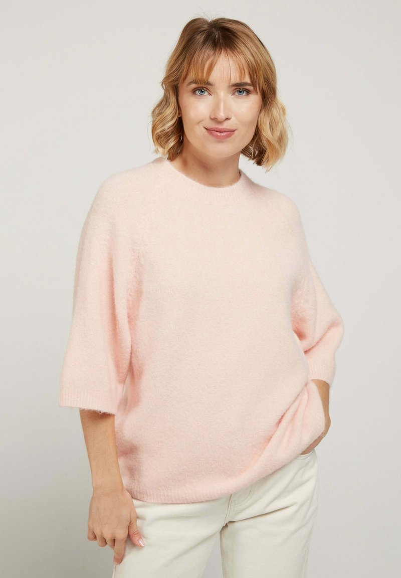 Un pull en tricot rose clair avec une coupe décontractée, des manches courtes et un col rond, fabriqué à partir d'un tissu doux et texturé.