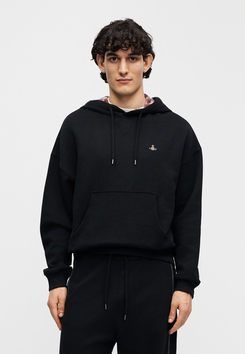 Sweat à capuche noir avec une poche kangourou, des cordons de serrage et un petit détail brodé. Porté sur une chemise à carreaux, accompagné d'un pantalon noir assorti.