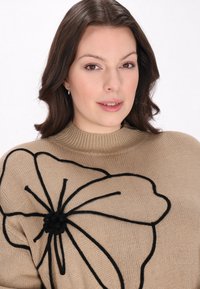 Pull en tricot beige arborant un grand motif floral noir et des accents texturés, avec un col à côtes haut et une coupe décontractée.