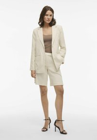 Blazer in lino beige con revers, abbinato a pantaloni corti coordinati. Top attillato marrone sotto e sandali con tacco alto neri e incrociati.
