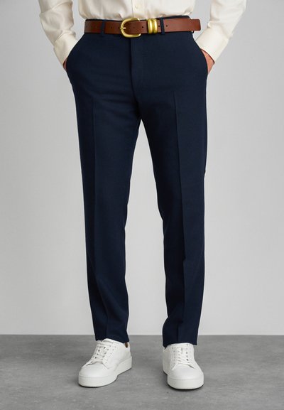Pantalones de marino con una textura suave, que presentan un corte ajustado, dos bolsillos laterales y un cinturón de cuero marrón con hebilla dorada. Combinados con zapatillas blancas.