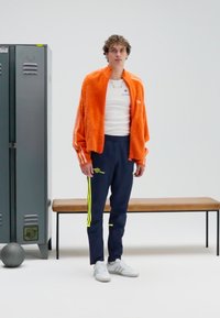 Pomarańczowa kurtka polarowa na białej koszuli, granatowe spodnie dresowe z żółtymi paskami oraz białe tenisówki. W tle znajduje się ławka i szara szafka.