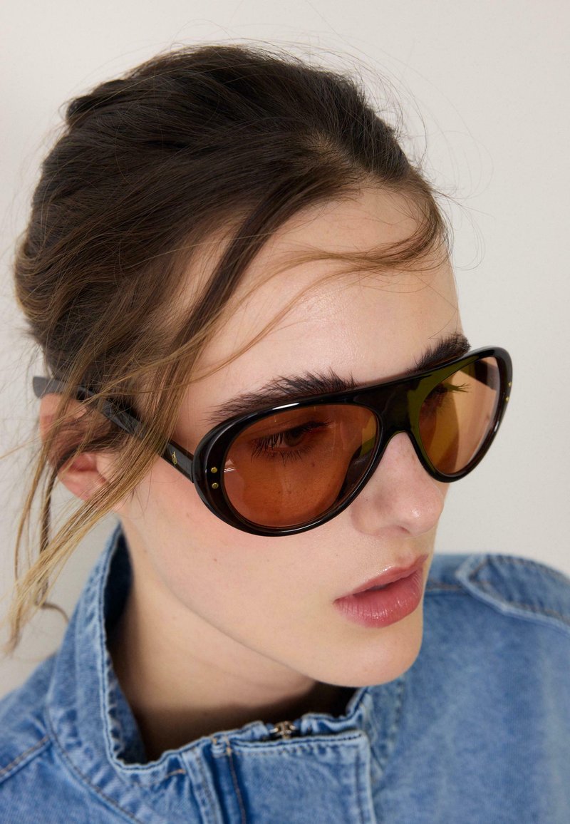Jeune femme portant des lunettes de soleil noires surdimensionnées avec des verres bruns et une veste en denim bleu, regardant légèrement vers le bas sur le côté.