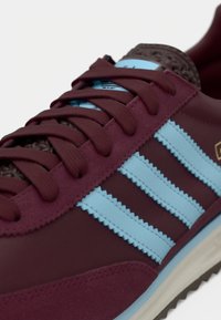 Bordo spalvos sportbačiai su šviesiai mėlynais zigzaginiais dryžiais, tekstūruotais rudais akcentais ir plokščiomis tamsiai raudonomis batų raišteliais. Turi šviesios spalvos guminį padažą.