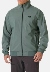 Chaqueta cortavientos en verde apagado, con cuello alto, cremallera frontal completa, puños elásticos y pequeño logo negro en el pecho. Textura suave.