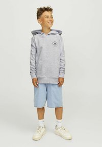 Hanorac gri deschis cu glugă, dotat cu un buzunar frontal și un logo circular negru, purtat împreună cu șorturi de denim albastre și adidași albi.