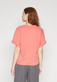Coral short-sleeve t-shirt with rolled cuffs, relaxed fit, soft fabric, and round neckline, worn with gray trousers.