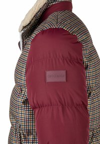 Close-up van een gewatteerde jas mouw met bordeauxrode stof en een geruit patroon, met een schapenwollen kraag en een "HECHTER" label op de arm.