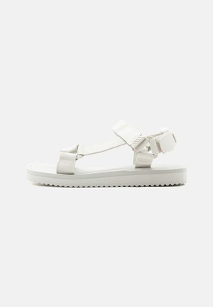 White flat sandal with adjustable ankle and toe straps and textured sole, designed for casual wear.