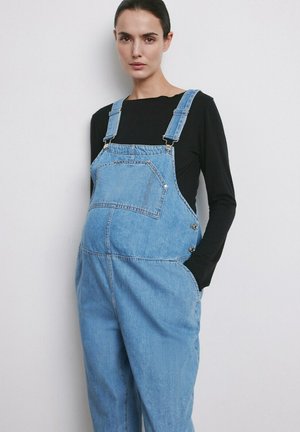 Mujer embarazada con peto de mezclilla azul claro y camisa negra de manga larga, de pie con una mano en el bolsillo frente a un fondo blanco liso.