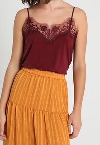 Canotta bordeaux con bordi in pizzo sovrapposta a una gonna maxi a righe arancioni. La parte superiore presenta spalline sottili e un delicato motivo floreale.