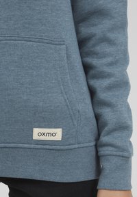 Blå sweatshirt med framficka, ribbad mudd och fåll, med en krämfärgad etikett med "oxmo" på den nedre sidan. Mjuk textur.