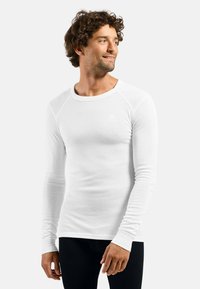 Long-sleeve white thermal shirt featuring a round neck, ribbed texture, and a small logo on the chest, paired with black pants.