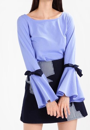 Femme portant un haut à manches longues violet clair avec des manches évasées nouées par des nœuds noirs, assorti d'une jupe noire ornée de détails blancs en patch.