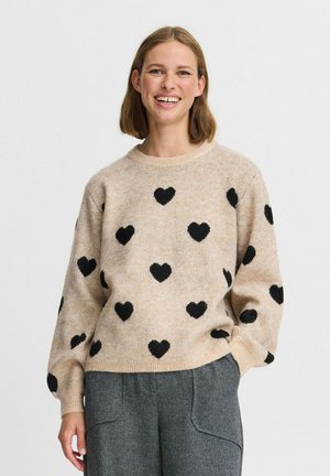 Motifs de cœurs noirs sur un pull en tricot beige clair avec un col rond et une coupe décontractée, associé à un pantalon gris. Tissu légèrement texturé.