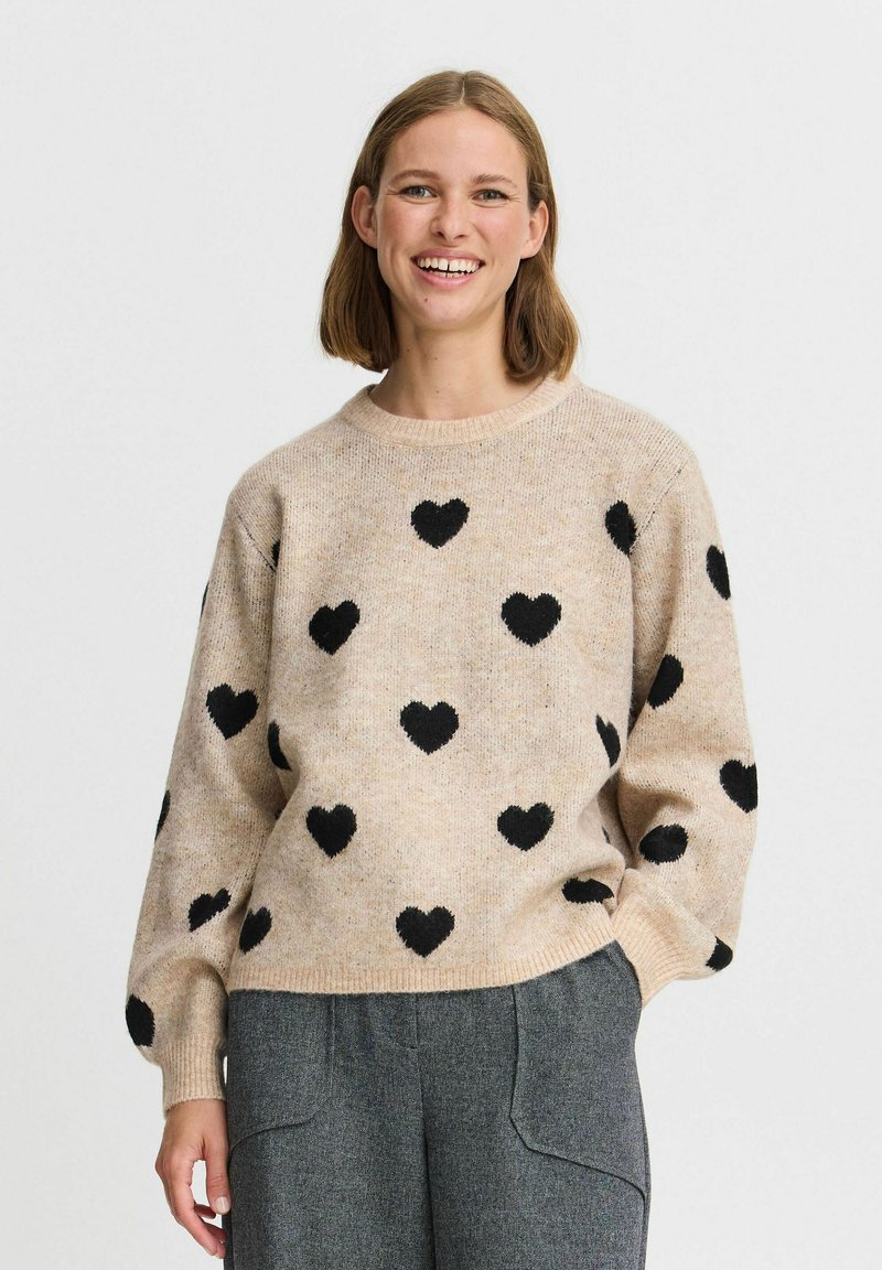 Schwarze Herzmuster auf einem hellbeigen Strickpullover mit Rundhalsausschnitt und entspannter Passform, kombiniert mit grauen Hosen. Leicht strukturierter Stoff.