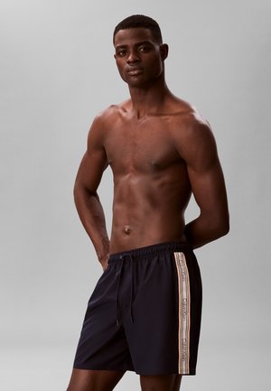 Uomo di pelle scura senza maglietta che indossa pantaloncini da bagno neri Calvin Klein con strisce laterali, in piedi davanti a uno sfondo grigio uniforme.