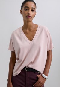 Jasnoróżowy t-shirt z dekoltem w serek, wykonany z miękkiej tkaniny, z krótkimi rękawami i luźnym krojem, zestawiony z czarnym paskiem i ciemnymi bordowymi spodniami.