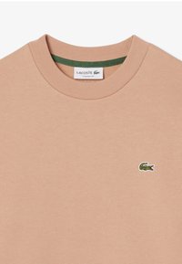 Camiseta de manga corta y cuello redondo en algodón color durazno claro con una textura suave. Presenta un logo verde de Lacoste bordado en el lado izquierdo del pecho.