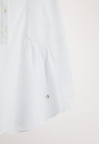 Valkoinen pusero napituksella, levenevällä helmaosalla ja koristeellisella hopeisella yksityiskohdalla alaosassa. Valmistettu kevyestä, hieman teksturoidusta kankaasta.