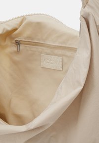 Interno di una borsa in tessuto beige con tasca con zip, etichetta testurizzata con testo in rilievo e rivestimento liscio. Design leggero e minimalista.