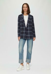 Marineblauwe double-breasted blazer met een lichtblauw geruit patroon, gedragen over een witte top, gecombineerd met lichtblauwe jeans met opgerolde mouwen en witte sneakers.