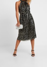 Žena v bezramínkových černých midi šatech se stříbrným vzorem, držící stříbrnou kabelku zdobenou kamínky, v černých lodičkách na kotníkové pásky.