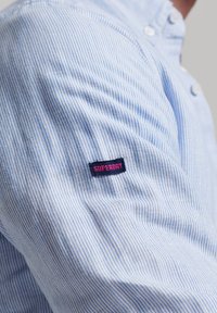Camisa a rayas azul y blanca con cuello botón y una tela texturada, que presenta un parche de logo "SUPERDRY" en negro y rosa en la manga.