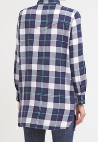 Navy blue and white plaid long-sleeve shirt with green accents, featuring a pointed collar, and a longer back hem, made of lightweight fabric.