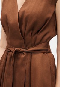 Καφέ φόρεμα χωρίς μανίκια με λαιμόκοψη σε στυλ wrap σε σχήμα V και υφασμάτινη ζώνη δεμένη σε κόμπο στη μέση.